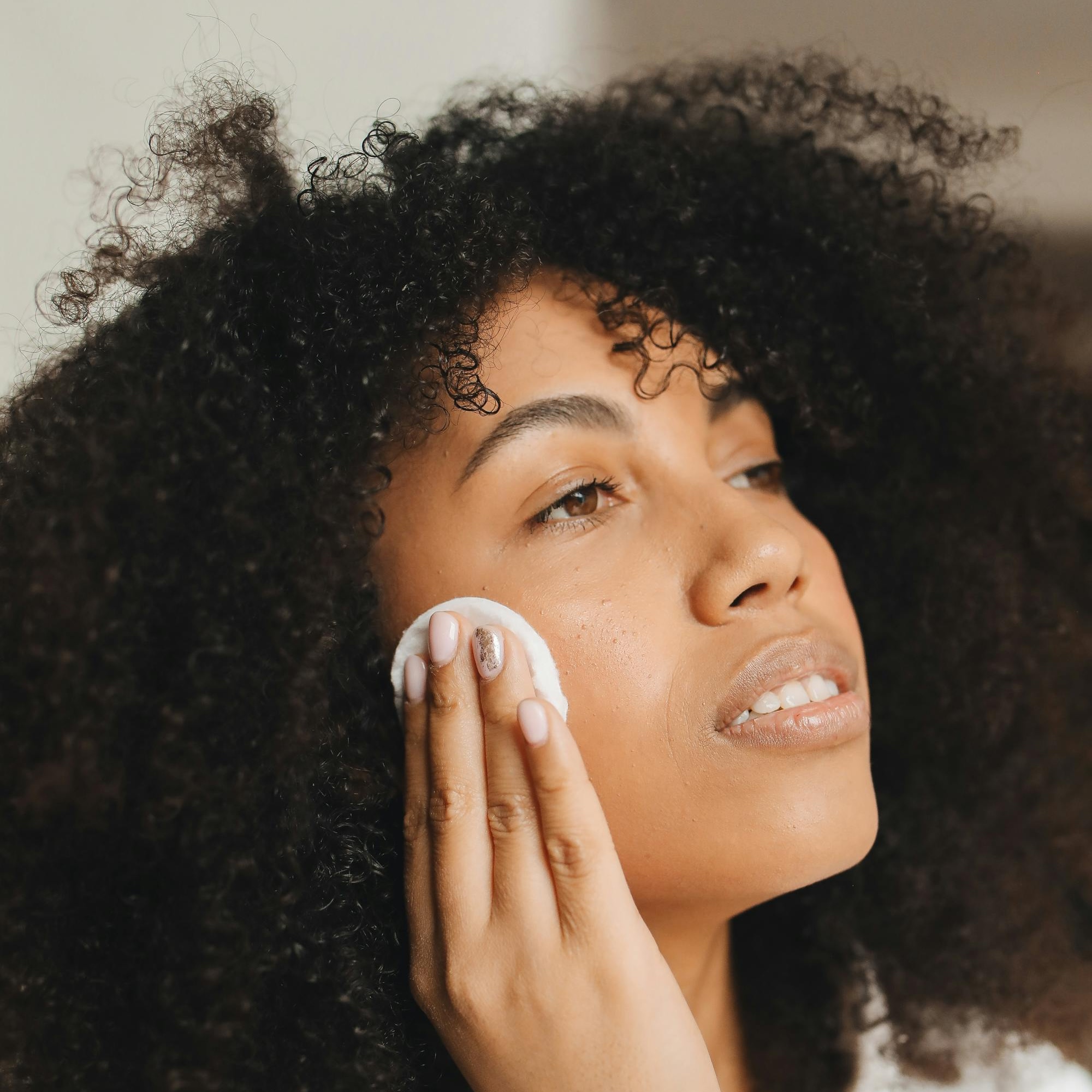 pexels-polina-tankilevitch-7382950 Frau mit lockigem Haar reinigt ihr Gesicht mit einem Wattepad, symbolisiert die Hautpflege und die Vorteile des Hydro Facials von RAU Cosmetics.